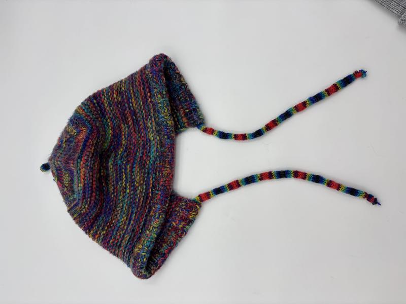 Handknit baby bonnet in multicolor variegated yarn with earflaps and braided tie strings, laid flat on white background