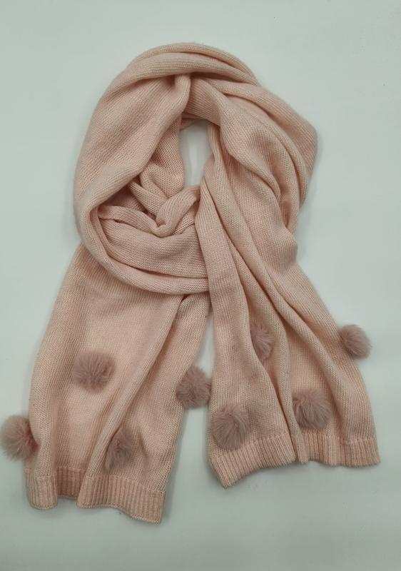 Soft blush pink knitted scarf with fluffy pom pom embellishments along the edges, loosely coiled on white background