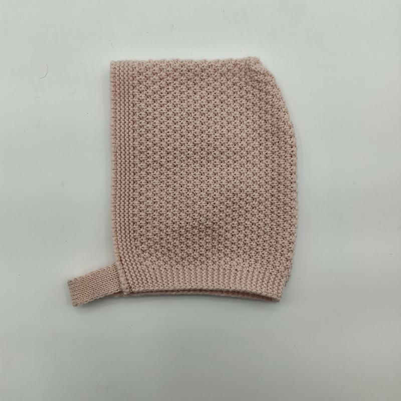 Flat lay of a soft blush pink knitted baby bonnet with seed stitch texture and ribbed chin strap on white background