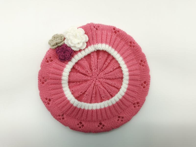 Pink knitted beret laid flat showing white circular stripe, eyelet lace pattern, and three small crochet flowers in white, mauve, and beige