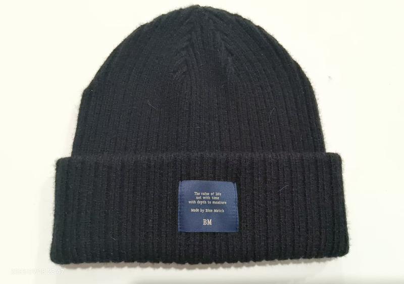 Black ribbed knit winter beanie hat with folded cuff and blue woven label patch on white background
