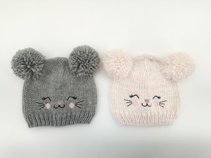 Two knitted baby beanies with pom-pom ears and embroidered cat faces, one gray and one light pink, on white background