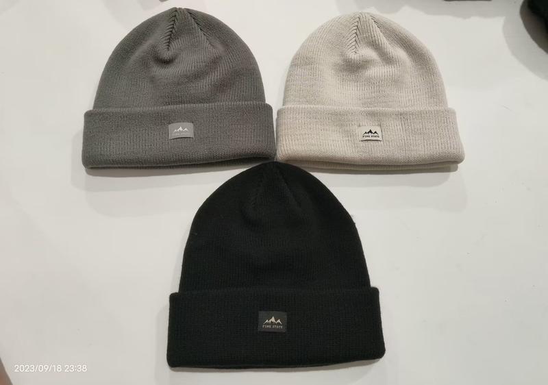 Three ribbed knit cuffed beanies in black, grey, and cream laid flat on white surface with mountain logo patch