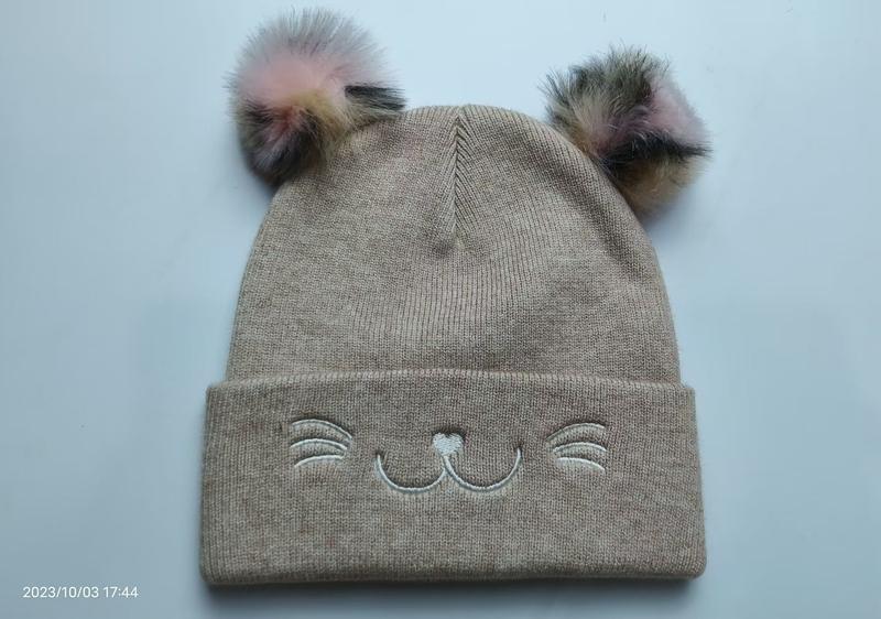 Beige knitted children's beanie hat with two multicolor fur pom poms and embroidered cat face design on cuff