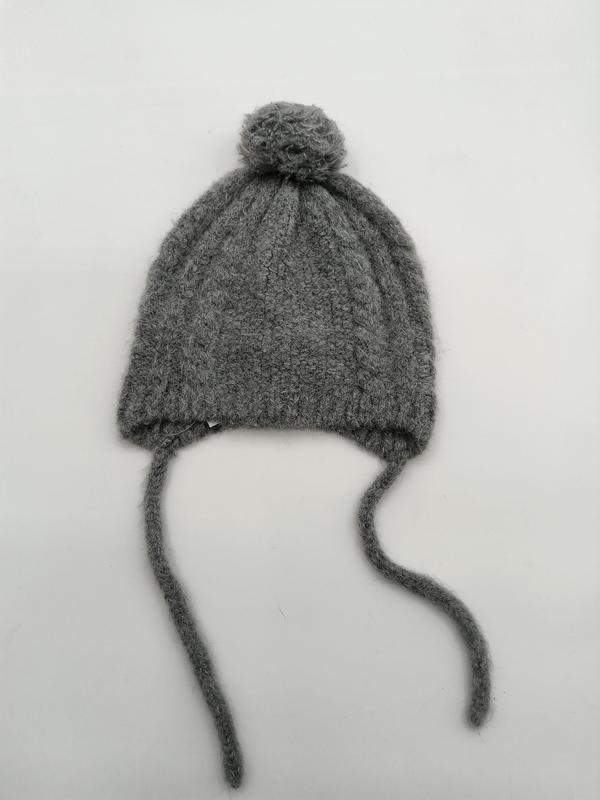 Gray cable knit baby winter hat with round pom pom on top and two long yarn tie strings on white background