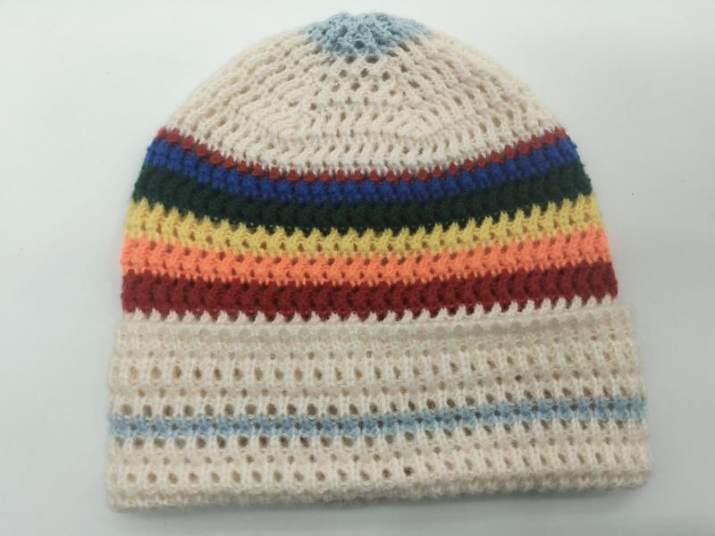 Handmade crochet beanie hat with rainbow multicolor stripes on cream open-weave background, laid flat on white surface