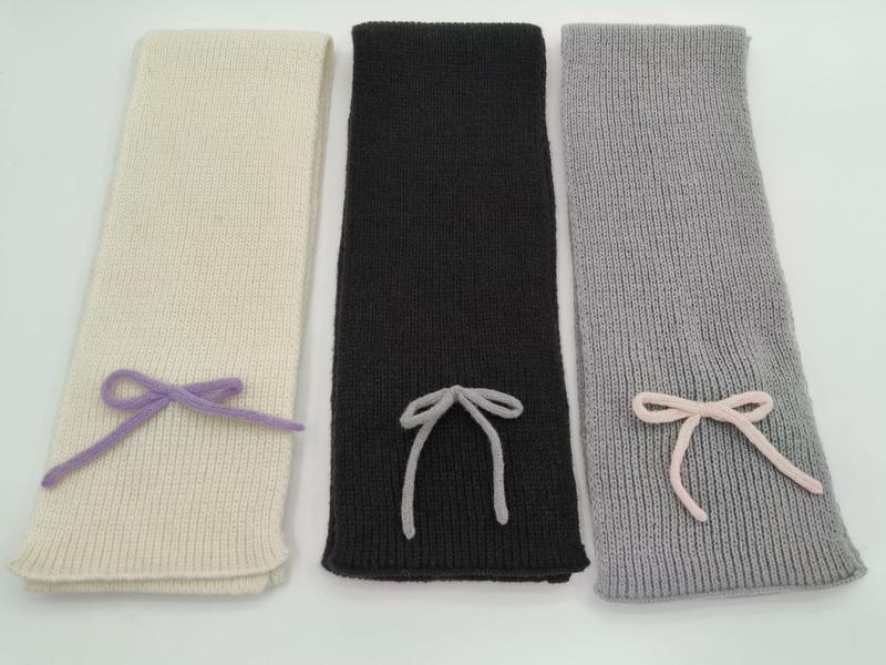 Three ribbed knit scarves in ivory, black, and grey, each with a small decorative yarn bow, laid flat on white background