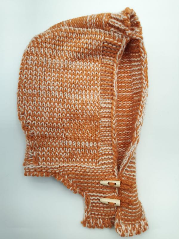 Hand-knitted baby bonnet in burnt orange and cream marled yarn with wooden toggle buttons on chin straps, laid flat on white background