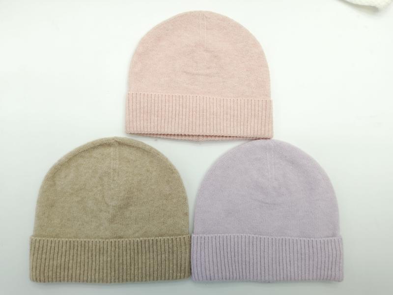 Three folded knit beanies in blush pink, taupe, and lavender with ribbed cuffs laid flat on white surface