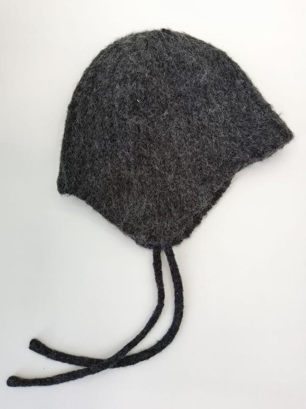 Handknit charcoal gray felted wool baby bonnet with ear flaps and braided tie strings laid flat on white background