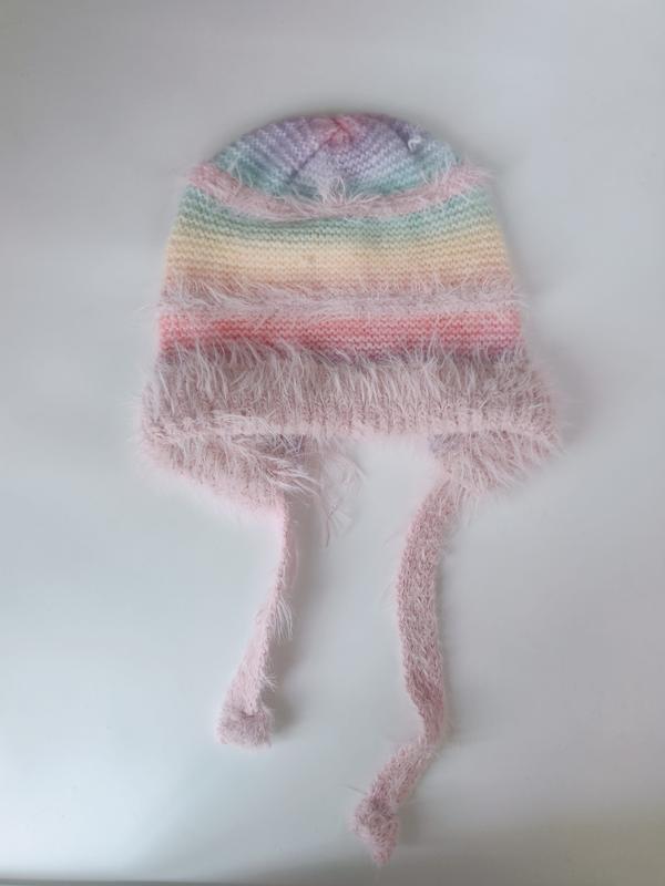 Handknit pastel rainbow striped baby beanie with fluffy pink eyelash yarn trim and long tie strings, laid flat on white surface