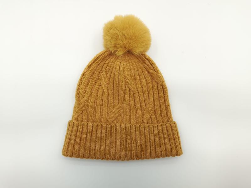 Mustard yellow cable knit winter beanie hat with large faux fur pom pom and ribbed cuff, laid flat on white background