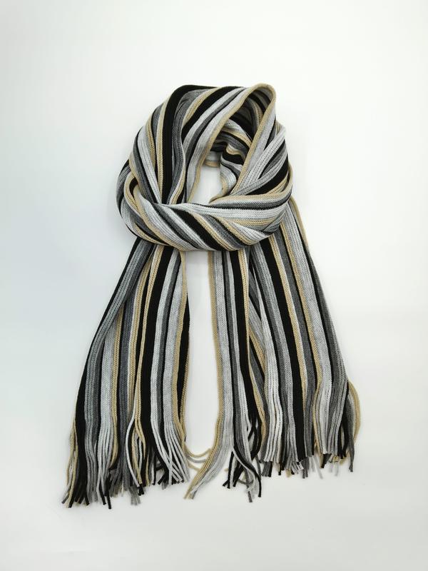 Knitted striped scarf in black, grey, and beige tones with tassel fringe ends, loosely looped on white background