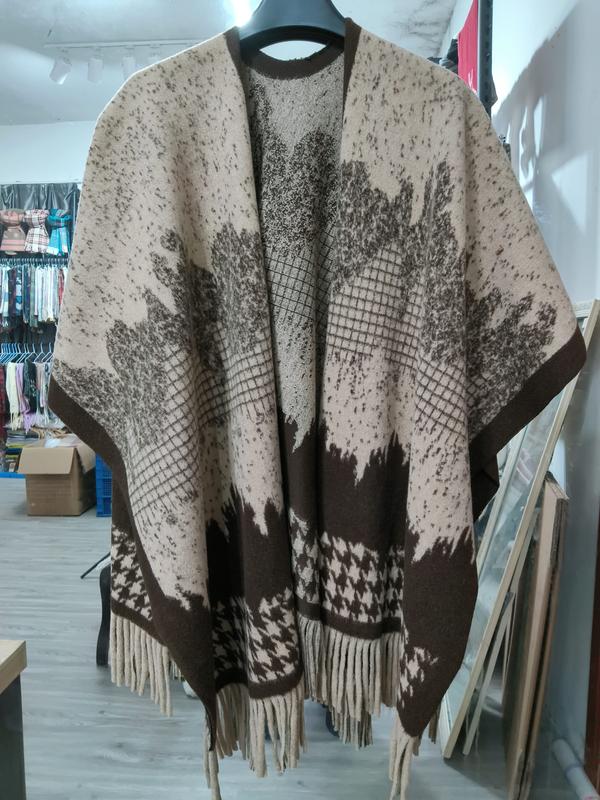 Oversized knit poncho cape in beige and brown with leaf print, houndstooth border, and fringe hem on hanger