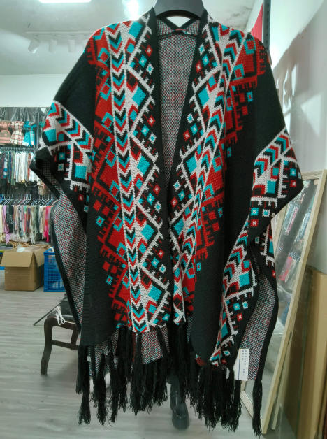 Black open-front knit poncho with red teal white Aztec geometric pattern and fringe hem displayed on hanger