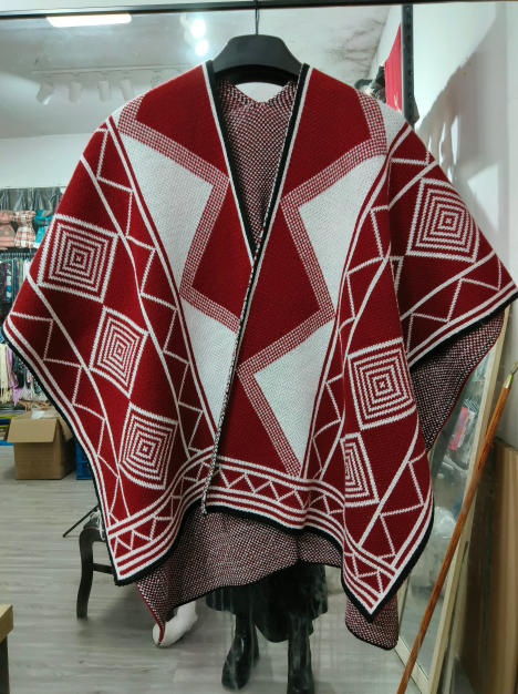 Red and white knitted poncho with bold geometric diamond and square patterns displayed on a hanger in a retail store