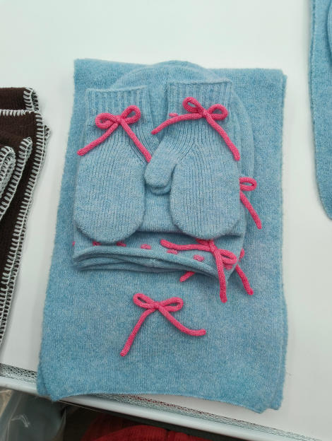 Baby blue knitted winter gift set including scarf, beanie hat and mittens with pink bow embellishments, displayed flat