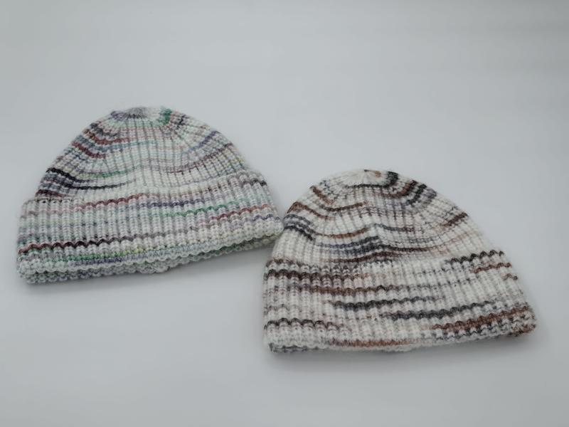 Two hand-knit ribbed beanie hats in white with multicolor stripes, one with cool tones and one with warm earth tones, on white background