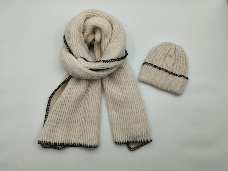 Cream ribbed knit scarf coiled next to matching beanie hat, both featuring black blanket stitch edge detailing on white background